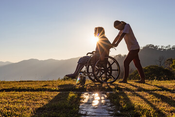 Disabled handicapped young man in wheelchair walking with his care helper in sunset.Silhouette