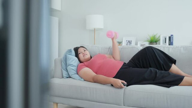 Asian funny oversize big girl use dumbbell workout while lying on sofa