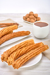 Spanish churros covered with sugar and cinnamon on a white plate accompanied by Spanish chocolate in a white cup on a marble surface and white background.