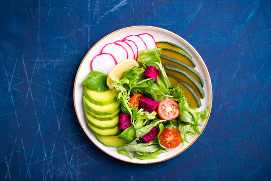 Healthy vegan salad fruits and vegetables dish with avocado, dragon fruit, radish, pumpkin, tomato, corn salad, lettuce, mizuna and lemon on blue background
