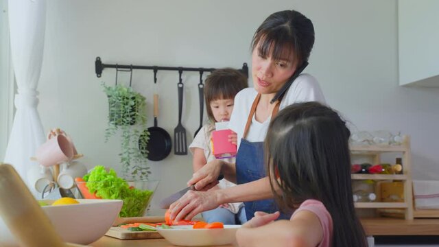 Asian busy mother talk on phone and cook with daughters in kitchen.