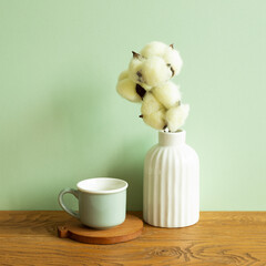 Mug cup and white vase of cotton plant on wooden table. green wall background. Home interior