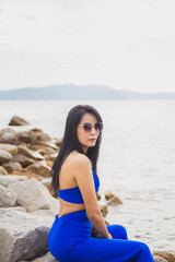 Fototapeta na wymiar Portrait Asian woman in blue casual wear with sunglasses sitting on the cliff above the sea while traveling in Asia