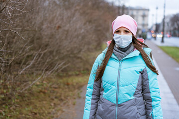 Fototapeta na wymiar a cute young woman of Caucasian ethnicity, in a warm jacket and a pink hat, in a medical mask, walks through the city streets in autumn. Cloudy day for a walk