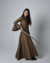 Full length  portrait of beautiful young asian woman with long hair wearing medieval fantasy gown costume. Graceful standing posing holding a sword,  isolated on studio background.