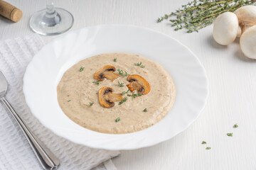 Fototapeta na wymiar Creamy mushroom soup puree made of blended champignon and dairy cream decorated with fried slices and thyme served for lunch on plate with spoon and glass of wine on white wooden table with textile