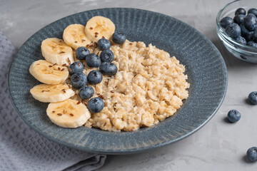 Oatmeal porridge made of boiled oats decorated with fresh blueberries and sliced banana served on blue gray plate on textile on concrete background for breakfast as source of protein. Horizontal