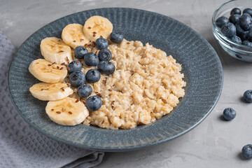 Oatmeal porridge made of boiled oats decorated with fresh blueberries and sliced banana served on blue gray plate on textile on concrete background for breakfast as source of protein. Horizontal
