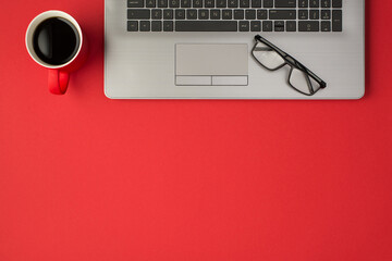 Top view photo of spectacles on grey laptop and red cup of coffee on isolated red background with copyspace