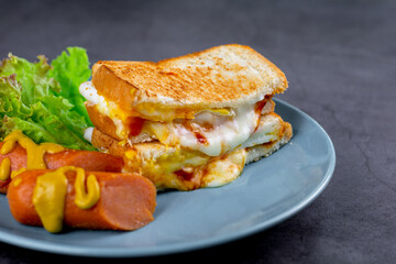 American breakfast concept. Sandwich with melting cheese and blurry sausage and fresh lettuce on a blue plate on a dark rock table. Homemade food in the morning.