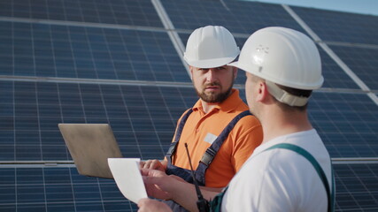 Close up engineers use laptop and project plan on the solar farm in special uniform standing look around discuss the installation of sunny batteries electricity environmental. Green energy jobs