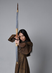 Close up  portrait of beautiful young asian woman with long hair wearing medieval fantasy gown. Graceful pose holding a long bow and arrow,  isolated on studio background.