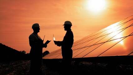 Two Unidentified Solar Power Engineers In Backlight At Sunset. Team of industrial colleagues using tablet monitoring rows of photovoltaic solar panels at sunset. Solar park. Alternative energy concept