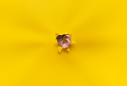 Close-up of a woman's ear through a torn hole in yellow paper. The concept of eavesdropping, espionage, gossip, tabloids and the yellow press. Background with copy space.