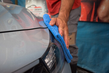 A hand cleaning the front of a gray car with a blue microfiber cloth, polishing the headlight with high shine in a car wash workshop