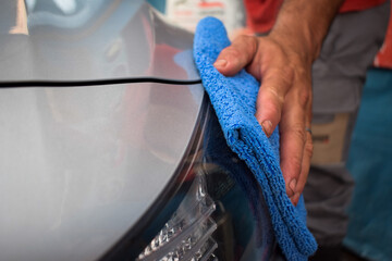 A hand cleaning the front of a gray car with a blue microfiber cloth, polishing the headlight with high shine in a car wash workshop