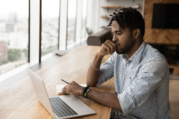 Pensive millennial African guy sit at table looks at laptop screen consider task, e-studying online...