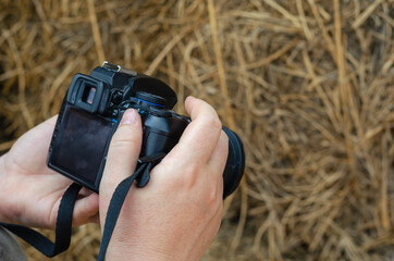 Woman is holding a black DSLR camera against dry yellow hay. A m - obrazy, fototapety, plakaty