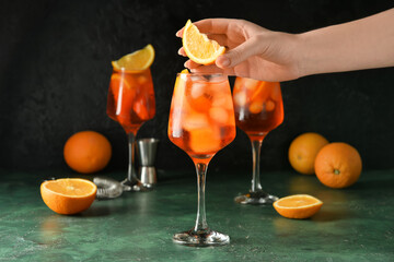 Woman decorating glass of tasty aperol spritz cocktail with orange on dark background, closeup