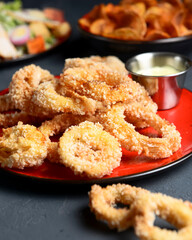 Golden onion rings and sauce in a saucepan served on a red plate. Over dark black background.