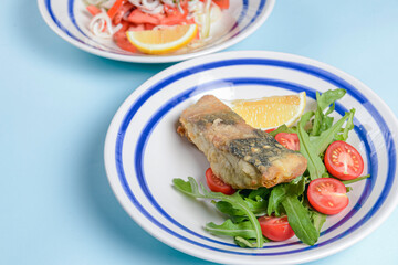 Delicious battered fish on a plate with cherry tomatoes, arugula and a piece of lemon.