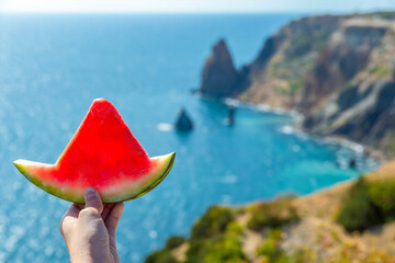 A slice of watermelon in the form of a boat on the background of the blue sea and picturesque coastal rocky mountains, located in soft focus.Summer beach concept. Diet of tropical fruits.To relax.
