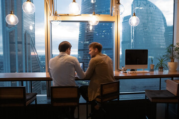 Back rear view two office employees male colleagues gathered in early morning solve urgent collaborative project sit at coworking table in modern office with city view. Teamwork, cooperation concept