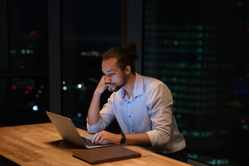 Busy 35s African ethnicity employee working hard using wireless computer stay late in modern skyscrapers office, night city view on background. Urgent online project, careerism, after hours concept