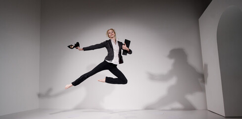 Graceful barefoot ballerina in a business suit jumping with shoes in her hands on a white...