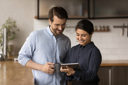 Diverse colleagues standing in kitchen office at lunch break discuss collaborative project use digital tablet. Married couple of business people talk in morning before workday, modern tech app concept