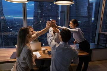 Multi ethnic successful colleagues finish meeting giving high five gesture. Group of young motivated ambitious startupper working until late in modern office, night city view through panoramic window
