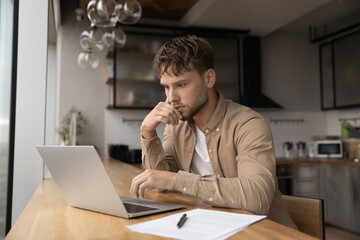 Thoughtful serious young 25s man student freelancer working or studying use laptop looks at device...