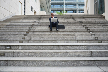 Pensive young businessman sitting on steps with opened laptop and working on important e-mail for business partner