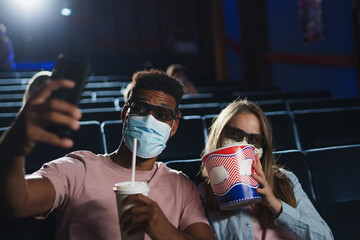Cheerful young couple with popcorn and 3d glasses taking selfie in the cinema, coronavirus concept.