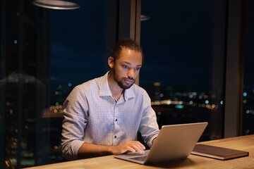 Busy 30s African male employee working hard using wireless computer stay late in modern skyscrapers office alone, behind his back night light city view. Urgent project, over hours, workaholism concept