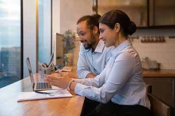 Diverse colleagues sit in office kitchen work use laptop, indian ethnicity mentor explain to...