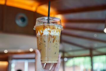 Iced caramel coffee. Hand holding a cup of coffee mixed with milk and caramel syrup at coffee cafe.