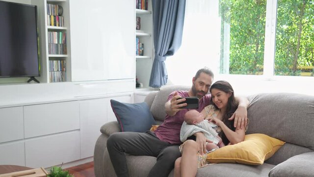 Caucasian husband use smartphone taking selfie or video call with his wife and sleeping newborn baby son in home living room. Happy family father and mother with infant child boy relationship concept