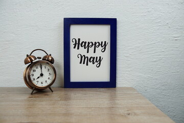 Happy May typography text with alarm clock and artificial plant decoration on wooden table and white wall and white wall background