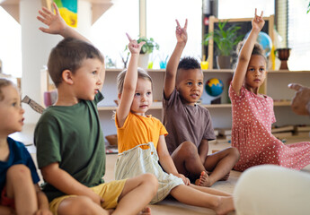 Group of small nursery school children sitting on floor indoors in classroom, raising hands. - obrazy, fototapety, plakaty