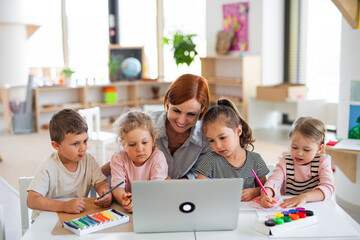 Group of small nursery school children with teacher indoors in classroom, using laptop.