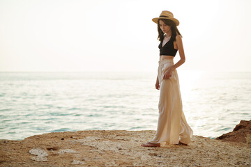Young stylish woman standing on cliff at the sea at sunset