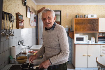 Portrait of elderly man cooking on stove indoors at home, stirring and looking at camera.
