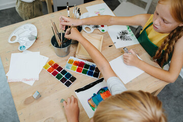 High angle image of kids painting with watercolor. They are keen and competitive, sitting behind the same small square table. Cleaning brushes at the same time.