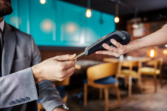 Hands holding a payment terminal. A man elegantly dressed in a business suit puts a credit or debit card in an online payment terminal held by a feminine hand. Paying bills for a meal at a restaurant