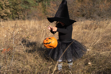 funny toddler girl in black witch hat with orange candy bucket in nature.
