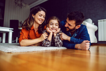Family lying down on stomach on the floor. Beloved daughter is laughing while parents showing her lots of love.