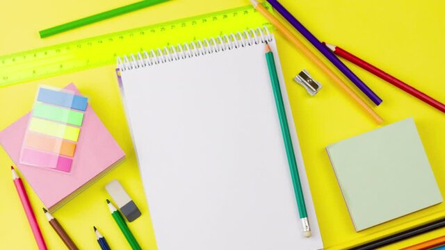 a white blank notepad lies on a pastel yellow background in the center with a variety of school supplies for school days. Concept for the first of September. Slow motion 4k, copy space