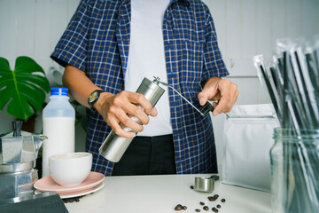 Barista is grinding coffee beans with manual stainless steel grinder to make black coffee machine, brewing equipment or coffee drip set Dripper on a wooden table In the kitchen at home in the morning