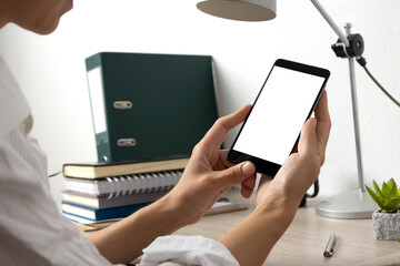 Closeup of blank white screen of mobilephone against office desk.Woman wearing white blouse and holding smartphone against stack of notebook,lamp and wooden table,white office wall